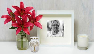 A small white urn with gold detailing on the mantle in front of an old woman's picture, flower vase, and a candle