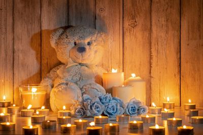A large teddy bear resting against a wooden wall, with light blue roses and a variety of candles placed around it.