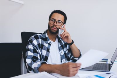 A man wearing glasses sits while talking on his phone  with a thoughtful gaze