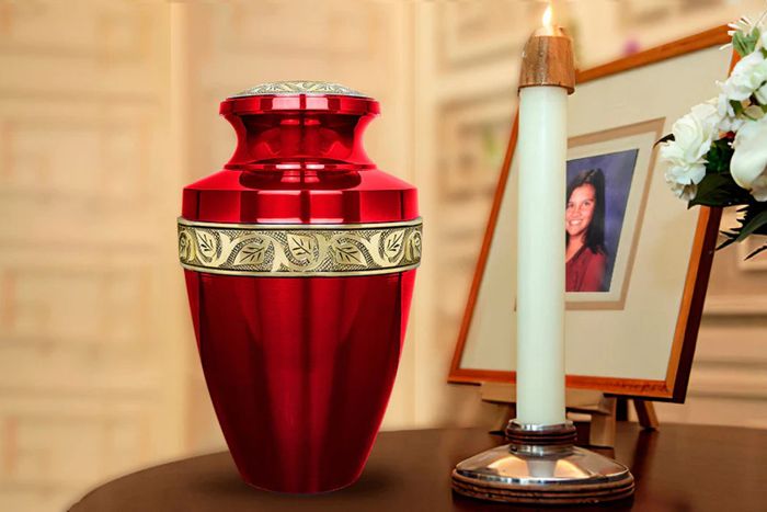 A red cremation urn with gold displayed on a table next to a candle and a picture of a woman preview image
