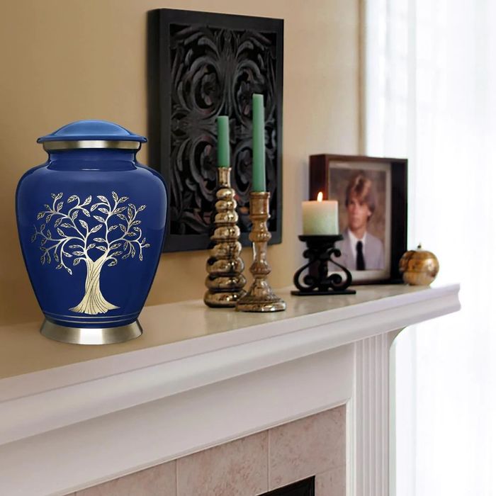 A dark blue urn with gold tree of life symbol on a mantle next to candles and a picture