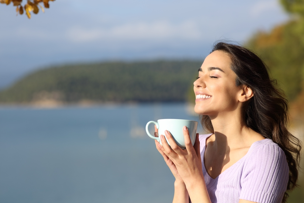 A woman smiling while holding a cup of coffee with a lake in the background