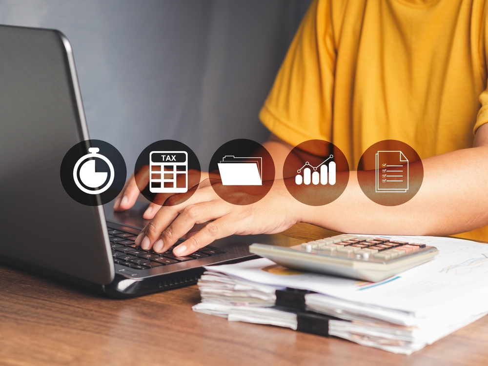 Woman sitting at a desk while working on a laptop with a pile of reports and a calculator next to it and an infographic of different payroll software features