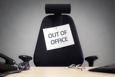 An office chair with a sign that reads "Out of office" behind a desk with a phone, glasses, computer mouse, and a keyboard