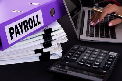 Close-up of a woman's hands typing on a laptop with payroll documents next to it