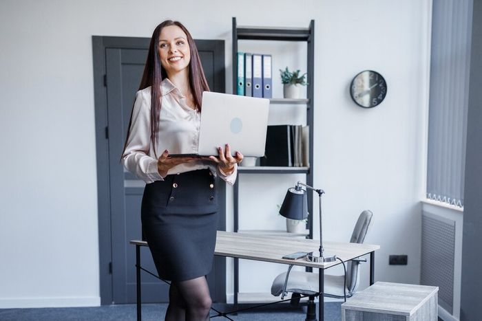 A young, professional woman with a pleasant demeanor, standing in an office holding a laptop.
