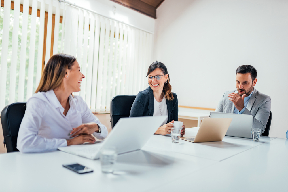 Three people sitting in an office with laptops talking and laughing