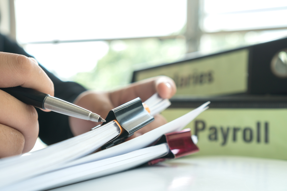 Close-up of a man writing in a notepad at an office desk with files marked "Payroll" and "Salaries" in the background