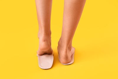 A person's feet placed on a pair of insoles, standing on their toes with the left foot, against a bright yellow background.