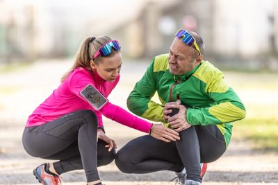 A woman assisting a man squatting holding his knee and grimacing in pain due to runner's knee
